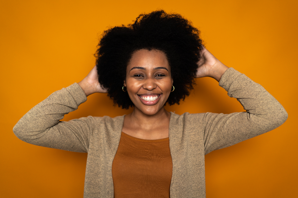 Black woman with natural curly hair smiling and touching her curls against a bright solid background, representing hormonal hair changes.